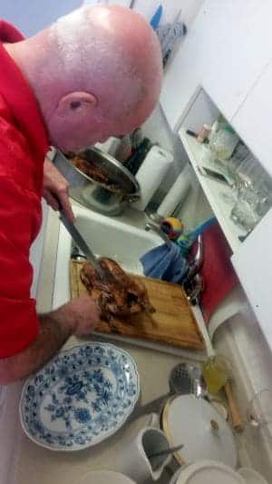 A man in a red shirt is carving a cooked chicken on a wooden cutting board during his road trip. A floral-patterned serving dish and various kitchen items are visible on the countertop, reminiscent of home comforts found along his journey.
