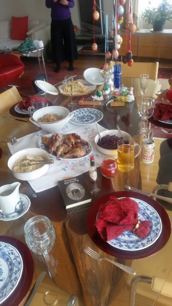 A dining table is set for a festive stop on your road trip, featuring a variety of dishes, colorful plates, and red napkins. Easter eggs hang from a display amid condiments and drinks. A person stands in the background, ready to continue the tour of delightful culinary experiences.