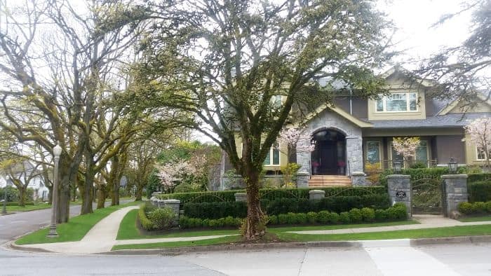 A suburban street with a large stone house surrounded by lush greenery and trees, perfect for a scenic road trip. The house boasts a neatly manicured front yard, and a sidewalk runs along the street. The overcast sky adds a calm atmosphere to the scene.