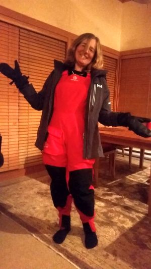 A person smiling and shrugging in a red outdoor suit with black accents, ready for a road trip. They're wearing black gloves and a dark jacket, standing indoors with wooden blinds and a dining table in the background.