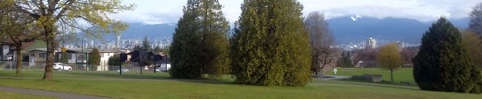 A scenic park view with green grass, trees, and a walking path invites a tranquil tour. In the background, a city skyline rises against snow-capped mountains under a cloudy sky, perfect for those dreaming of their next road trip adventure.