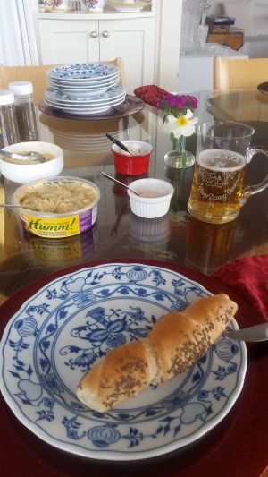 A dining table set for a road trip feast features a blue and white patterned plate with a bread roll, alongside a glass mug of beer, a decorative bowl, and tin of hummus. In the background, stacked plates and a small flower vase add to the cozy atmosphere.