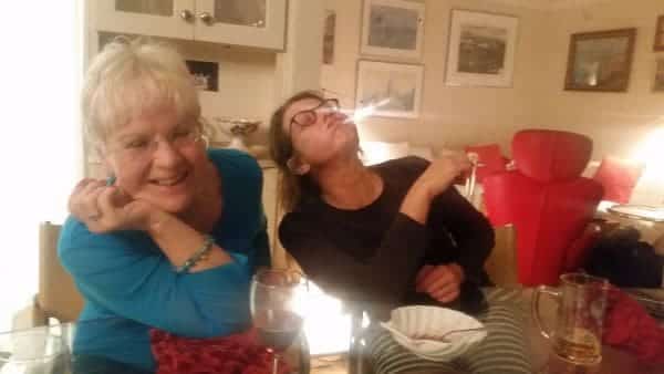 Two women sit at a table in a cozy room adorned with framed pictures. The woman on the left smiles, resting her chin on her hand, while the woman on the right, wearing glasses and reminiscing about their unforgettable road trip, playfully blows smoke.