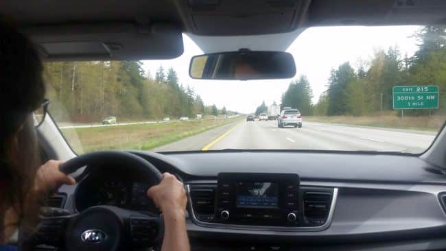 View from the interior of a car showing the driver’s hands on the steering wheel during a road trip. The vehicle is traveling on a highway with cars ahead. A green sign indicates Exit 215 and 300th St NW. Surrounding trees line the road under an overcast sky, setting the tone for an adventurous journey.