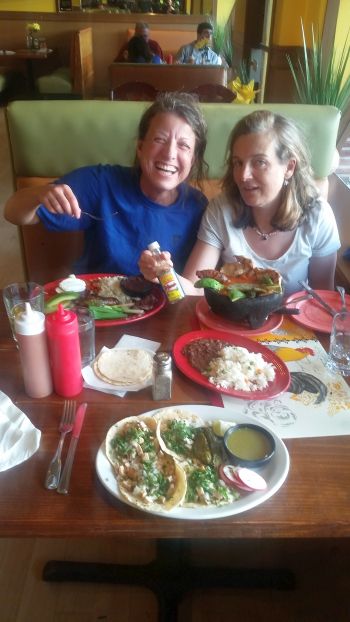 Two people on a road trip sit at a restaurant table, enjoying a meal. The table is filled with plates of food, including tacos, rice, beans, and hot sauce. One person is smiling broadly, holding a fork, while the other is eagerly eyeing the feast.