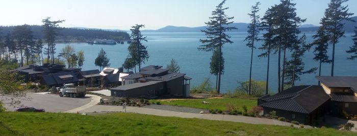 Experience the scenic view of modern houses by a serene blue body of water, perfect for your next road trip. Surrounded by towering evergreen trees, the area offers breathtaking mountains visible in the distance under a clear sky.