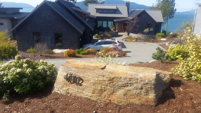 A modern house with dark wooden siding overlooks a scenic body of water. In the foreground, a large stone displays the address. The landscaped driveway curves up to the house, perfect for a road trip stop, with a white car parked near the entrance.