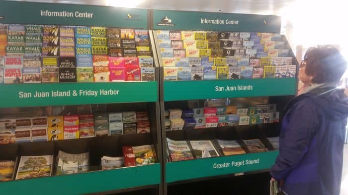 A person stands in front of brochure racks labeled "San Juan Island & Friday Harbor," "San Juan Islands," and "Greater Puget Sound" at an information center, planning a road trip. The racks are filled with colorful brochures that promise exciting tours and adventures.