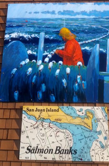 A vibrant mural depicts a fisherman in a yellow hat and red jacket skillfully working with nets on a boat. Below, a chart reveals water depths and locations labeled "San Juan Island" and "Salmon Banks," making it an intriguing stop on any road trip or tour of coastal treasures.