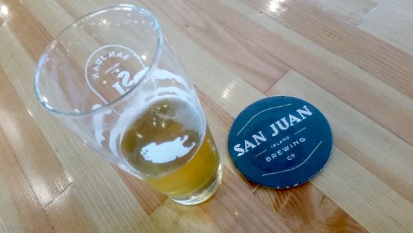A half-full beer glass sits on a wooden table next to a round coaster that reads "SAN JUAN BREWING Co." The glass is adorned with foam and features a white logo, making it the perfect pitstop for your road trip exploration.
