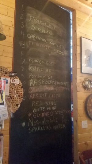 A chalkboard menu on a wooden wall lists various drinks, perfect for a road trip refreshment: English ale, blonde, caramel amber, stout, mango gose, kolsch, porter, raspberry wheat, apricot cider. Wines and non-alcoholic options like sparkling water complete the tour of flavors.