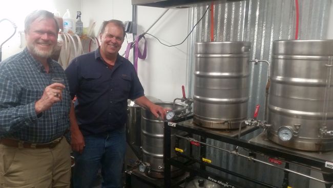 Two men are standing in a brewery room on their road trip. One holds a glass, while the other points at brewing equipment made from repurposed stainless steel kegs. Both are smiling, surrounded by corrugated metal walls and various pipes as they enjoy their impromptu tour.