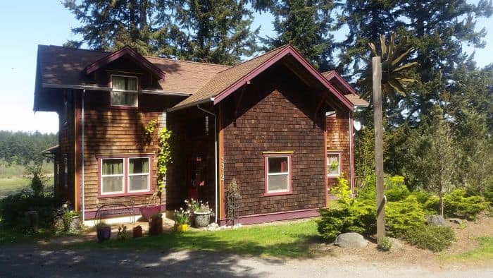 A rustic wooden house with red trim sits serenely in a grassy area surrounded by tall trees. Perfect for a road trip detour, the house features plants and pots near the entrance, a gravel path in front, all under a clear, sunny sky.