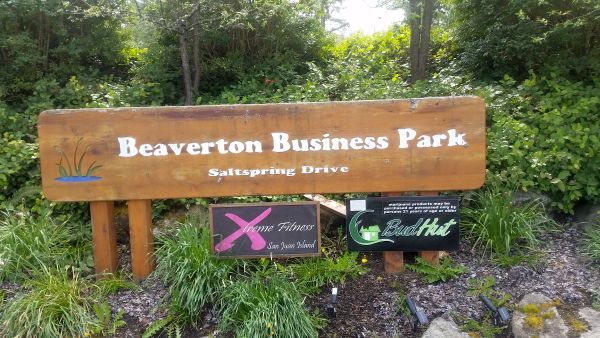 A wooden sign reads "Beaverton Business Park, Saltspring Drive," perfect for a road trip discovery, with smaller signs for "Xtreme Fitness" and "Bud Hut." Surrounded by lush green plants and trees, it sets the tone for your tour of local gems.