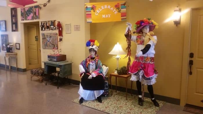 Two mannequins dressed in colorful traditional outfits are displayed in a room, capturing the vibrant spirit of a festive road trip. One is seated next to a lamp, while the other stands holding a ribbon. A banner above them reads "Celebrate May," and the room is adorned with vibrant accents.
