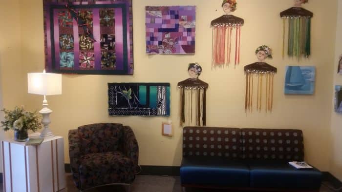 A cozy room invites you to pause your road trip, with its patterned armchair, small sofa, and white lamp on the side table. The walls showcase colorful art pieces, including patchwork quilts and fringe elements. A vase of flowers adds charm to this welcoming rest stop.