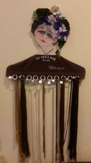 Decorative hair accessory display shaped like a hanger with a painted face and flowers, perfect for showcasing during your next road trip. Various brown hair extensions hang from ring clips at the bottom, adding style and flair to any travel adventure.