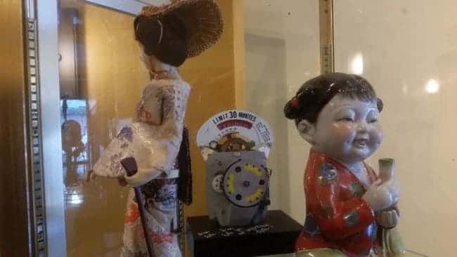A display case on our tour showcases two traditional-style dolls: one, an adult in a kimono with intricately styled hair; the other, a smiling child holding an object. A clock is visible in the background, marking time on this unique road trip through cultural history.