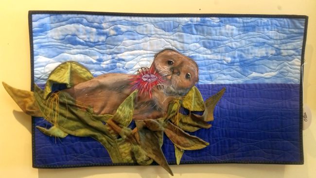 Quilt art depicting a sea otter floating in blue water under a cloudy sky invites you on a visual road trip. The otter holds a red sea urchin among green kelp leaves, while textured stitching adds realistic charm to this fabric masterpiece.
