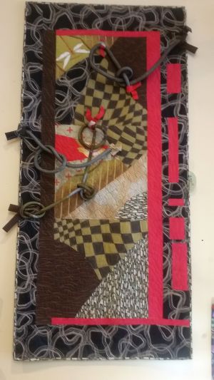 A colorful abstract textile artwork reminiscent of a road trip's vibrant spirit, featuring checkered patterns, swirls, and a mix of brown, red, and beige hues. The piece is adorned with rope knots and framed by a bold red border.