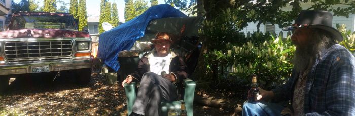 Two men sit outdoors in lawn chairs under a tree, one holding a drink. A vintage red truck, ready for a road trip, is parked nearby, partially covered by a blue tarp. Tall trees and a white house are visible in the background.