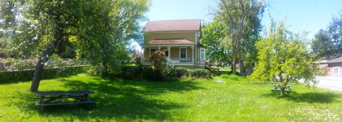 A charming two-story house with a brown roof, perfect for a road trip detour, nestled in a lush green yard. Tall trees provide shade over a wooden picnic table on the left and a smaller table to the right. The sky is clear and blue, hinting at an invitingly sunny day.