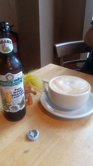 A bottle of Samuel Smith's Organic Pale Ale and a ceramic cup of cappuccino sit on a wooden table, evoking memories of a scenic road trip. A troll doll with yellow hair stands next to the bottle, its quirky charm adding to the scene. Nearby, a bottle cap completes this quaint tableau.