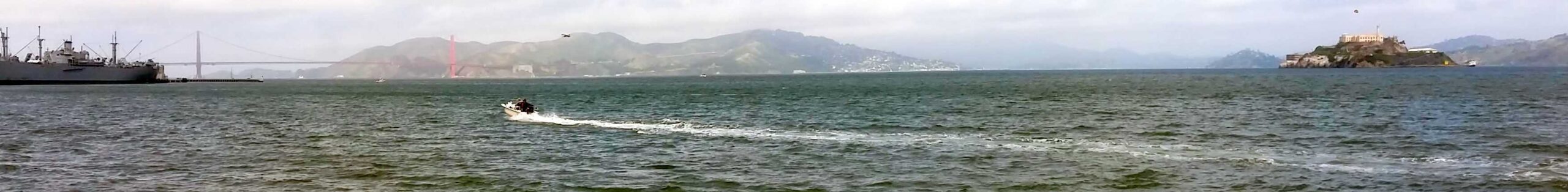 A small motorboat speeds across the water, leaving a white wake. In the background, the Golden Gate Bridge spans the bay, while a hillside and Alcatraz Island are visible under a cloudy sky—a perfect scene for an unforgettable tour of San Francisco's iconic sights.