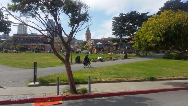 In a picturesque park setting, people lounge on benches beside a grassy field, while a cyclist tours along the path. Trees and buildings frame the scene under a partly cloudy sky.