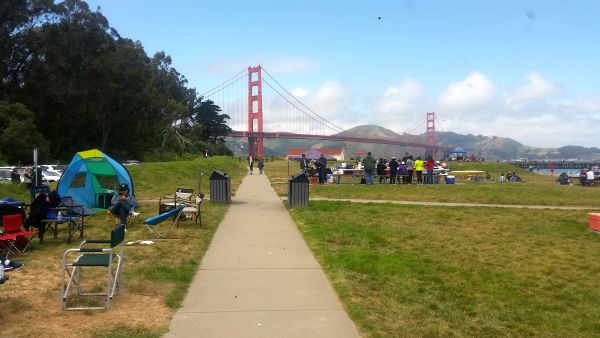A scenic view of a park with the Golden Gate Bridge in the background sets the stage for picnics and road trips. People are enjoying themselves near a few tents, under a partly cloudy sky. The lush green grass invites all to relax and savor the moment.