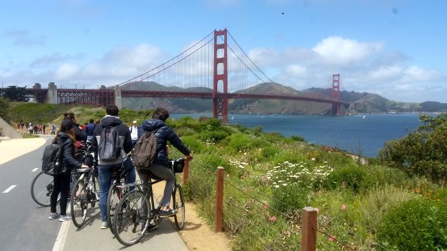 A group of cyclists on a road trip pause on a path overlooking the Golden Gate Bridge. The bridge spans across the blue waters with hills in the background. Wildflowers and greenery border the trail under a partly cloudy sky.