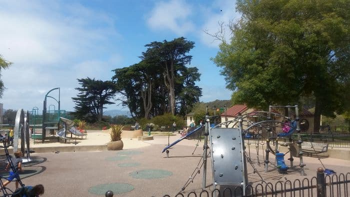 A playground with various climbing structures, slides, and swings is nestled among trees and fences. The bright blue sky dotted with clouds paints a perfect backdrop. Hills rise in the distance, reminiscent of a familiar stop on a memorable road trip where children play joyfully.