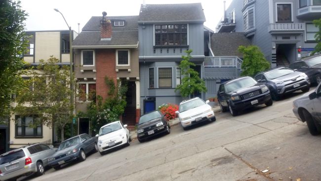 Cars parked on a steep, slanted street in a residential area with houses in the background offer a unique stop on your road trip. The vehicles appear tilted due to the incline, while diverse architectural styles of homes are nestled among lush greenery.