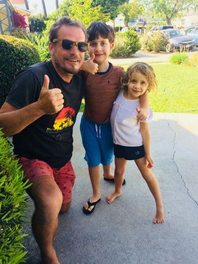 A man in sunglasses kneels on the driveway beside two smiling children, capturing moments from their road trip. The boy in the middle sports a brown shirt and blue shorts, while the girl wears a light pink top with navy shorts. The man gives a thumbs-up against a backdrop of greenery and yard bliss.