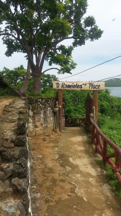 A stone path leads to a wooden sign reading "D' Romielea's Place" in a lush, green environment. Trees and foliage surround the entrance, with a glimpse of water hinting at nearby island hopping adventures. The area has a serene and natural ambiance.
