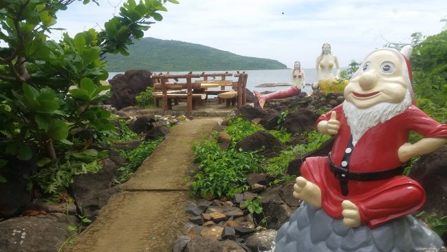 A garden path leads to a wooden table and bench by the sea, surrounded by rocks and greenery. A garden gnome statue sits atop a rock in the foreground. Nearby, a colorful kite surfing sail dances in the breeze, while two mermaid figures watch from the background with a hill across the water.