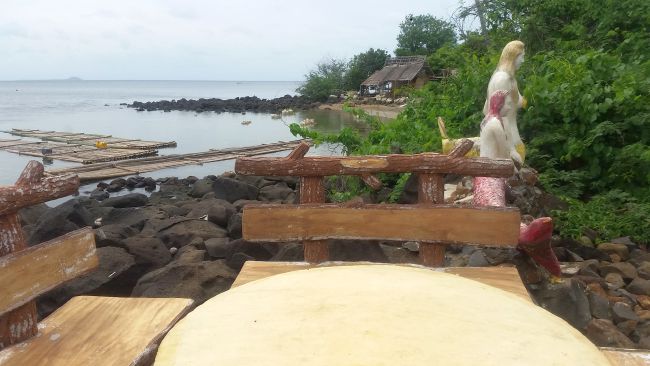 A serene coastal view features a round wooden table and benches in the foreground. A weathered statue stands amidst lush greenery by the water's edge. Bamboo rafts float nearby, hinting at an island hopping adventure, with a rustic hut visible in the background under a cloudy sky.