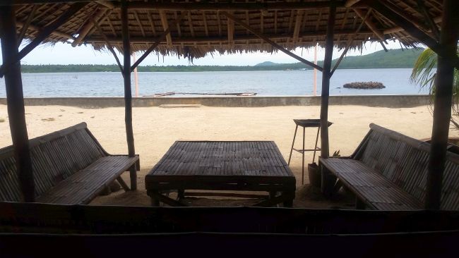 A shaded seating area with wooden benches and a table under a thatched roof overlooks a sandy beach, perfect for island hopping or kite surfing. The calm ocean stretches out to small islands and a mountain visible on the horizon, offering an idyllic view of Cuyo's serene beauty.