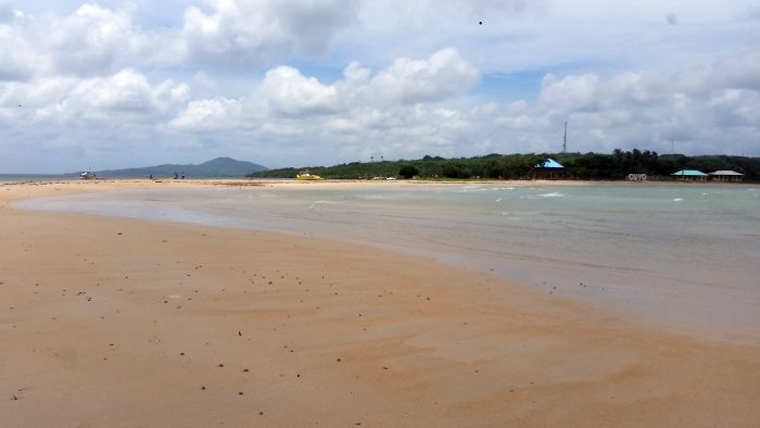 A serene beach scene unfolds with a wide stretch of light brown sand and gentle waves, perfect for Cuyo island hopping. Sparse clouds dot a blue sky, and small hills with greenery rise behind. A few small buildings are nestled along the shoreline, completing the picturesque setting.