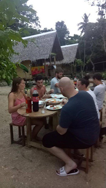 A group of people sits around a wooden table outdoors in El Nido, enjoying a meal and drinks. Tropical huts and lush greenery paint the backdrop, hinting at adventures like boat tours to nearby islands and a rustic yet relaxed setting.