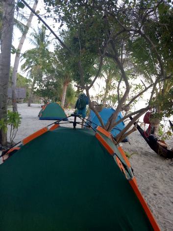 A row of colorful tents set up on a sandy beach in Coron, Palawan, shaded by palm trees. People relax in hammocks and stroll around, enjoying the various accommodation options. The ocean is not visible but implied by the setting. The atmosphere is peaceful and tropical.