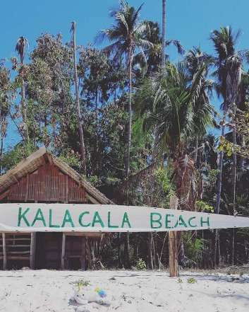 A bamboo hut stands behind a white sand beach with a "Kalacala Beach" sign, offering charming accommodation options. Tall palm trees rise under a clear blue sky, creating a tropical atmosphere reminiscent of El Nido's allure.