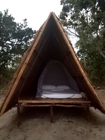 A rustic, triangular bamboo shelter set among trees in Palawan, with an elevated platform containing a bed covered by a mosquito net. The structure is open at one end, showcasing its simple, natural design—a perfect escape after exploring El Nido's stunning boat tours.