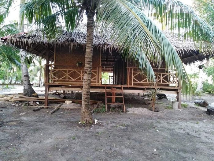A rustic, elevated wooden hut in Palawan with a thatched roof is nestled among palm trees. The hut features a railed porch and steps leading up to it. The sandy ground and lush tropical vegetation make it a perfect retreat after exploring boat tours around Coron.