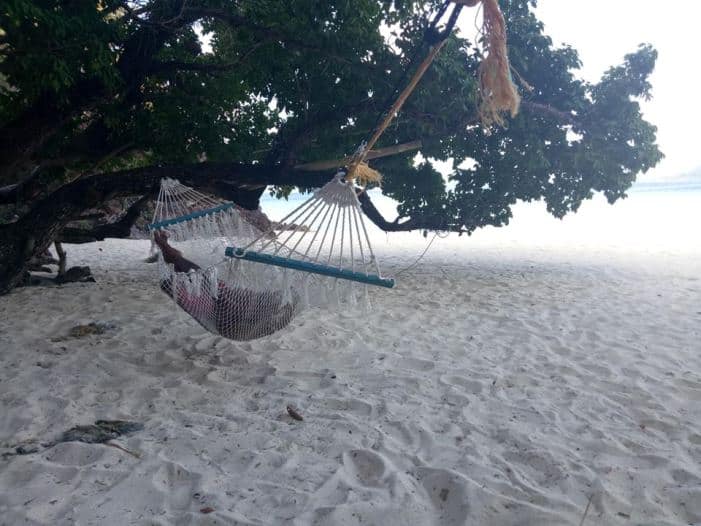 A person relaxes in a hammock tied to a tree on a sandy beach in Coron, Palawan. The ocean is visible in the distance, and the tree provides shade from the sun. This idyllic scene complements the area’s charming accommodation options for a perfect getaway.