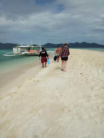 Three people stroll along a sandy beach in Coron, Palawan, heading toward a traditional outrigger boat docked on the shoreline. The calm sea and distant mountains under a partly cloudy sky set a serene backdrop for potential boat tours.
