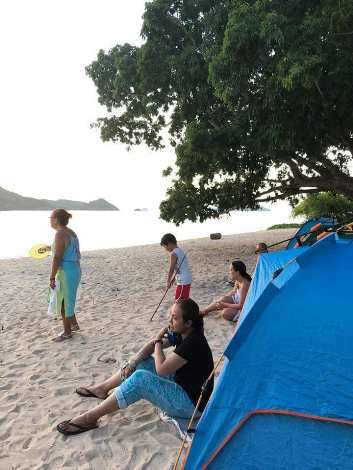 A group of people relaxes next to a blue tent on a sandy beach in Palawan. A child plays with a stick nearby, while a large tree provides shade. The ocean is calm in the background, with a distant island visible under the clear sky—perfect for boat tours exploring Coron.