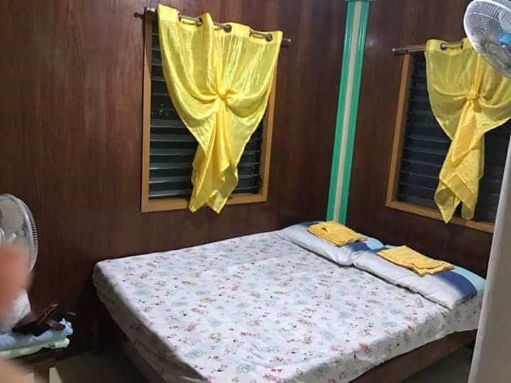 A cozy bedroom with wooden walls offers a charming accommodation option, featuring a double bed covered in a floral sheet. Bright yellow curtains adorn two windows, and a fan is visible in the corner. Two yellow towels are neatly placed at the head of the bed.