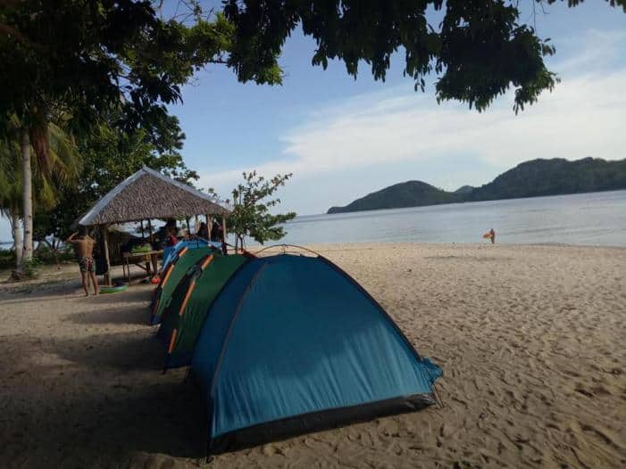 A row of blue tents is set up on a sandy beach under the shade of trees near El Nido. Nearby, a small thatched-roof shelter stands, and in the background, the calm sea meets distant green hills under a partly cloudy sky—a perfect spot for exploring local accommodation options or enjoying boat tours.