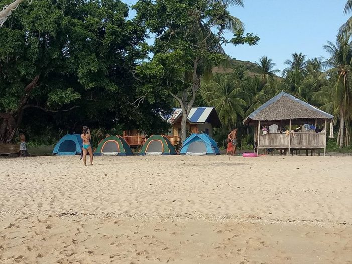 A sandy beach with several tents set up in front of a small, thatched hut in El Nido. People are walking nearby, enjoying the lush backdrop of green trees and palm trees. It's the perfect spot to start your Palawan adventure or embark on one of the famous boat tours.